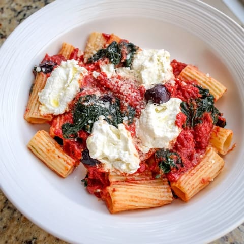 A close-up of a bubbling baked ziti, bursting with olives, spinach, and a cheesy top.
